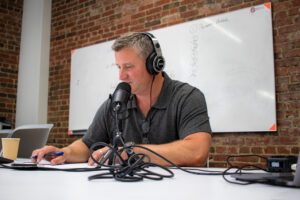 Spot Co-founder Andrew Elsener sitting at a white desk, wearing headphones, and speaking into a podcast microphone to record an episode of the More Than a Broker podcast. Speaking on Transportation Law