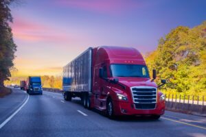 Red semi-truck hauling a trailer down a highway with other trucks following, representing long-haul freight transportation and truckload logistics on an open road.