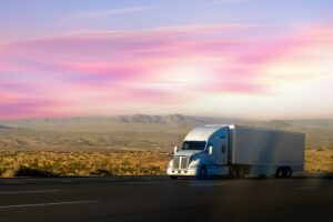 White semi-truck hauling a trailer across an open highway through a desert landscape at sunset, representing long-haul freight transportation and national supply chain logistics.