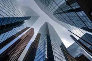 A low-angle, worm's-eye view looking up at a cluster of modern glass skyscrapers against a cloudy sky.