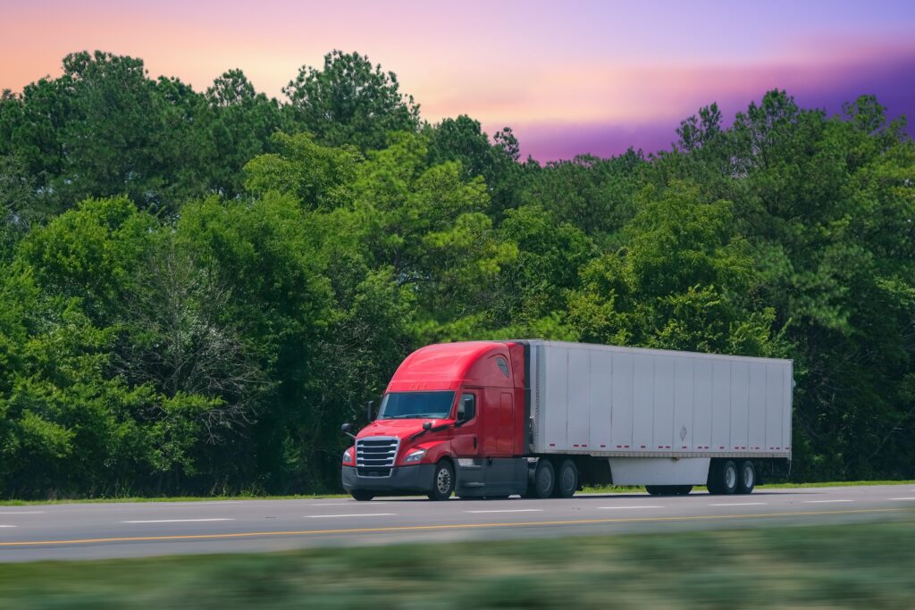 Red semi truck with a white trailer, alone on a road with green trees and a pink and purple sky in the background