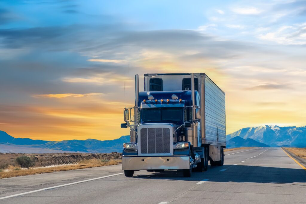Large blue semi truck driving on an empty road, sunset in the background