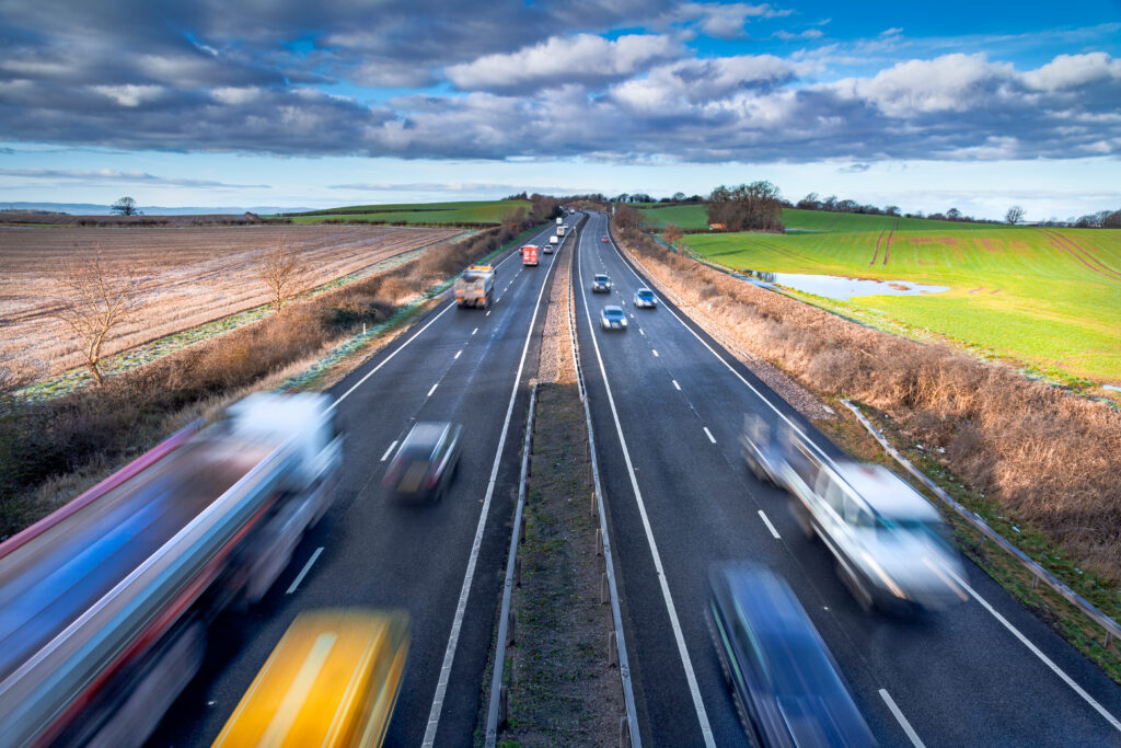 An elevated view of a multi-lane highway with blurred cars and trucks driving through a green rural landscape under a blue sky.