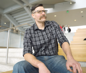 Gentleman with glasses and large mustache sitting on bleachers.